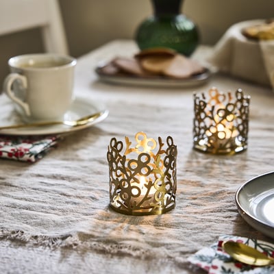 Golden VINTERFINT tea light holder, stainless steel with acrylic coating, intricate mistletoe design, small size, on tablecloth among other items.