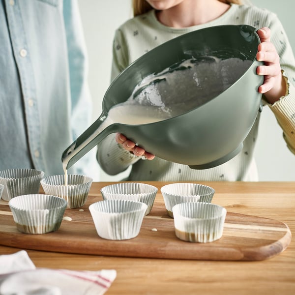 Person pouring batter from green VISPNING mixing bowl with tilted handle into cupcake liners on wooden board.