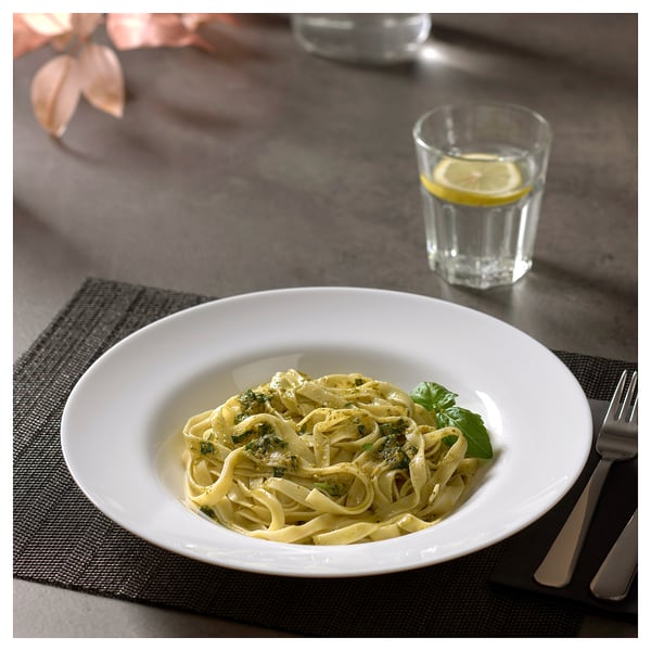 White fettuccine with pesto in bowl, water with lemon slice, fork and knife on mat.