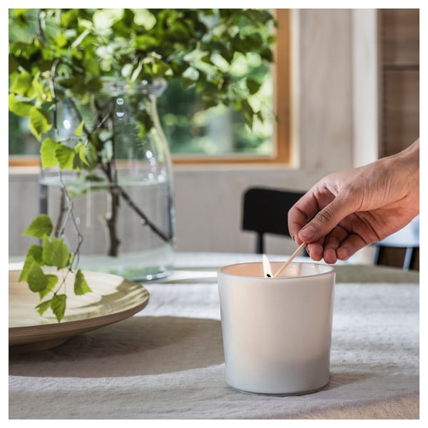 In a hand is lighting an ADLAD scented candle. The white ceramic jar sits on a light-colored cloth next to a vase with green branches.