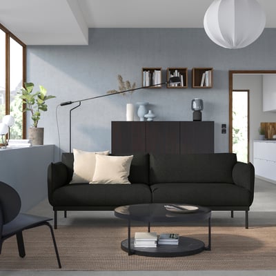 Modern living room: dark grey sofa, beige pillows, black coffee table, grey rug, blue vase, large windows, plant, bookshelf.
