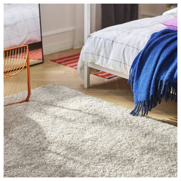 A bedroom with a white bed covered by a blue blanket, a red and white striped rug, and a fluffy grey rug on the floor.