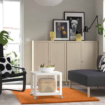 Modern living room with orange rug, white table, grey sofa, black-white chair, wood cabinet, & framed pictures.