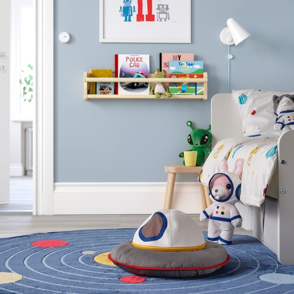 Soft spaceship toy in grey and red, made of recycled polyester. Perfect for pretend play. Placed near a small bed, indicating a childs room setting.