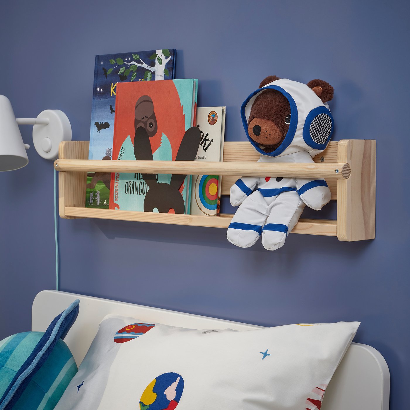AFTONSPARV astronaut teddy on shelf with books, beside white bed, blue wall. White & blue, soft toy, small.