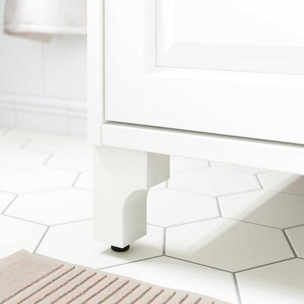 White wooden cabinet legs with black adjustable feet, set on hexagonal white tiles.
