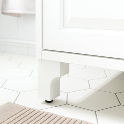 White wooden cabinet legs with black adjustable feet, set on hexagonal white tiles.