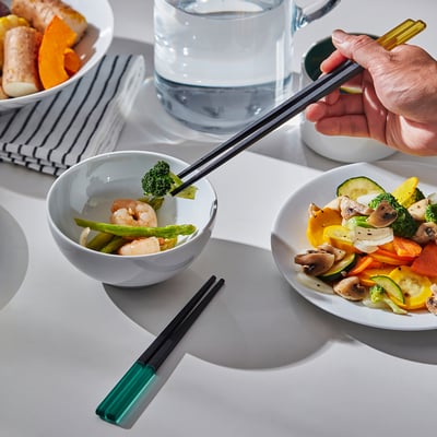 Hand holds coloured chopsticks near bowls of veggies and shrimp. Square tips prevent rolling.