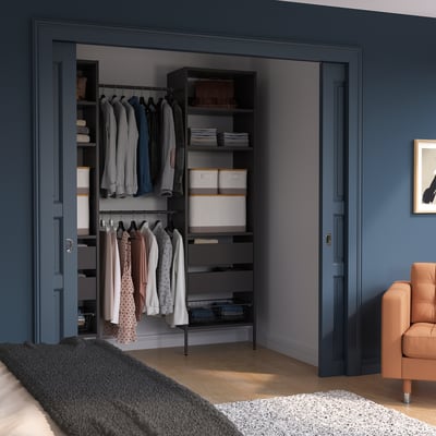 Organised closet with dark blue AURDAL storage. Hangs and shelves for clothes. Complementary furniture includes a beige chair and black bed.