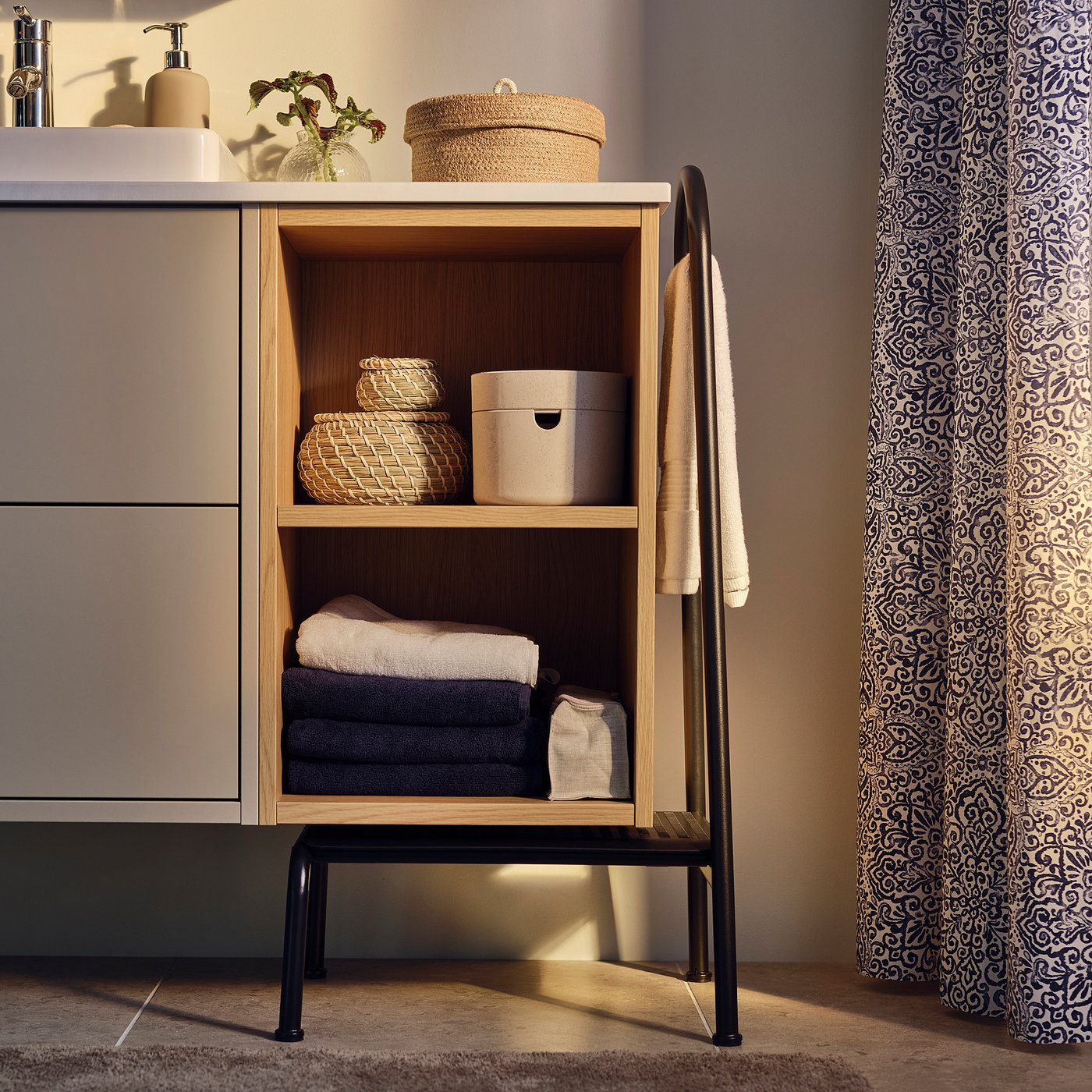 Modern bathroom with light wood BÄSINGEN vanity, towels, baskets, and patterned shower curtain.
