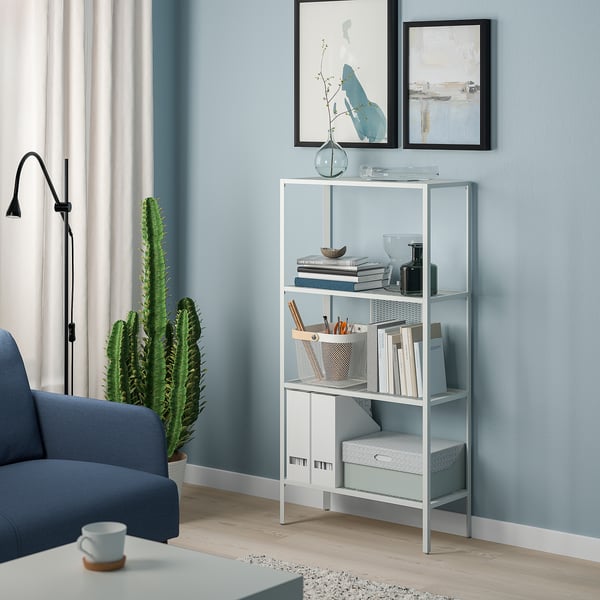 Modern silver metal BAGGEBO shelf with books and vases against blue wall. Large cactus and blue chair nearby.