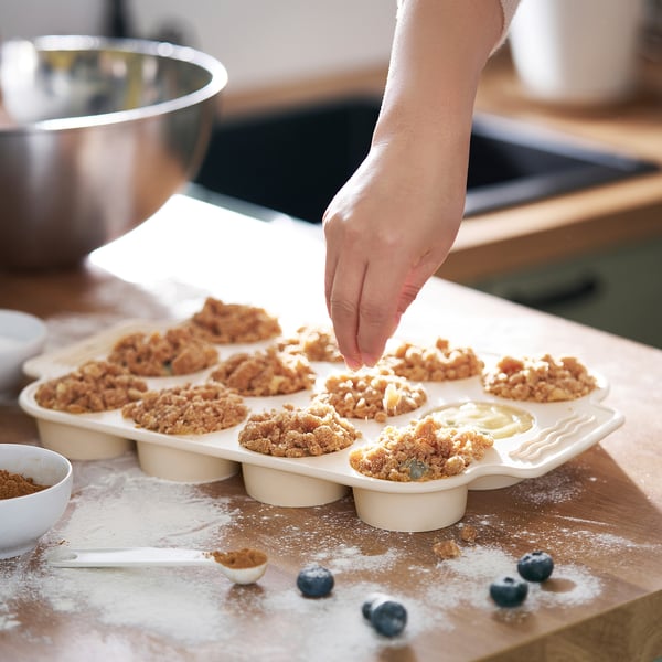 Hand placing topping on muffins in a 12-cup silicone baking pan on a floured surface.