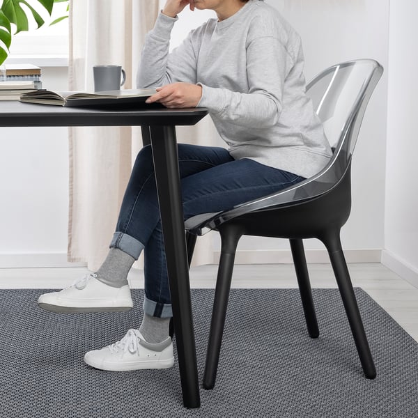 Person sitting at modern desk. Black chair with transparent seat. On black desk, grey items and cup seen.