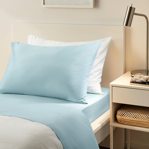 Blue bedding set on white bed, flat & fitted sheets, pillows, beside table with lamp, basket.