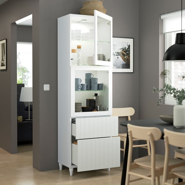 Modern kitchen with white BESTÅ cabinet displaying dishware, featuring glass doors, open shelf with cups, and two drawers. Wooden chairs seen nearby.