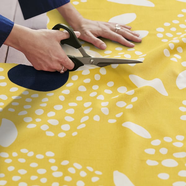 A pair of hands holding scissors cuts a bright yellow fabric with white floral patterns, likely preparing it for sewing.