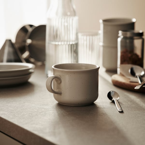 White speckled mug sits on table beside spoon, stack of plates, coffee jar, and container.