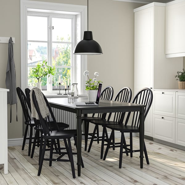 Small, modern black dining set fits cosy near large window. Six chairs surround rectangular table, hanging lamp above. White kitchen background.