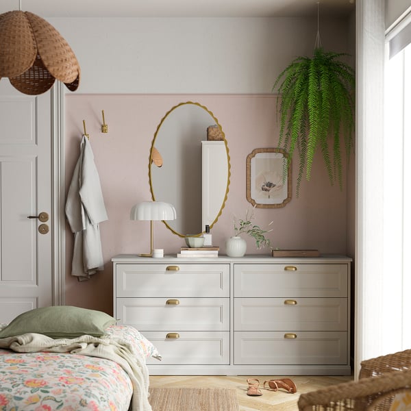 Pastel bedroom with pink wall, brass dresser, gold mirror, hanging plants, cosy bed.