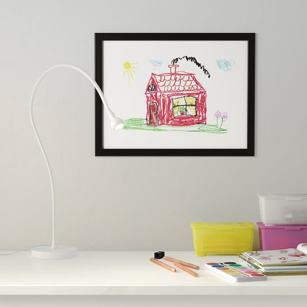 A childrens art frame hangs on a wall, displaying a drawing of a red house. On the desk is a white lamp, colourful storage boxes, pencils, and paint.