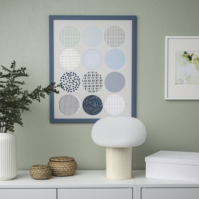 Modern gallery wall with blue-framed abstract art, white lamp, plant, and baskets.