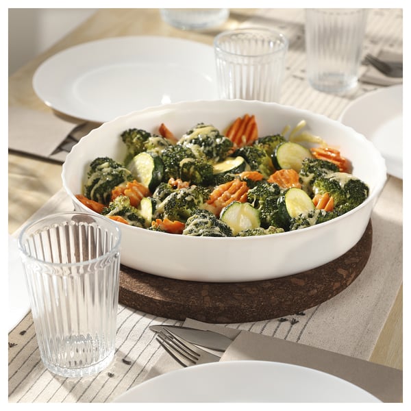 A glass dish with broccoli, carrots, and cheese on a table. Two clear glasses and plates surround it.