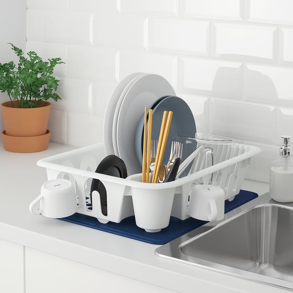 White dish drainer, FLUNDRA, holds plates, bowls, cups, cutlery. Placed on blue mat near sink, with plant nearby, drying dishes efficiently.