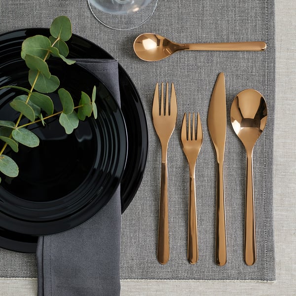 Modern table setting with black plate, grey napkin, gold cutlery, and green plant on grey placemat.