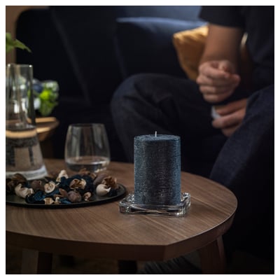 Grey candle in glass holder on wooden table, with wine glass and stones. Blurred person in background.