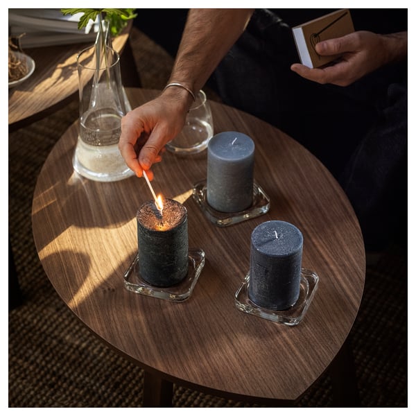 Hands light candles on wooden table, one with book, other with lighter. Three grey candles in glass holders, sunlight and vase.