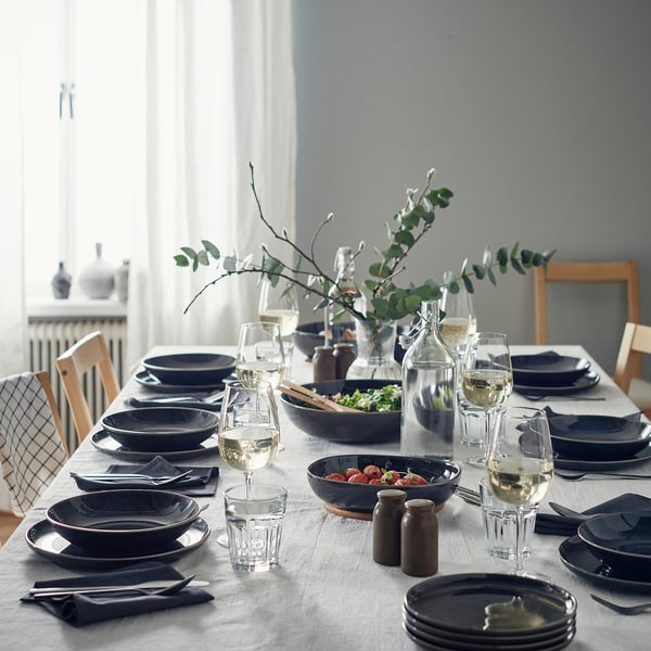 Elegant dining setup with GLADELIG dark-grey deep plates, clear glasses, neatly folded napkins, and a central vase with greenery.