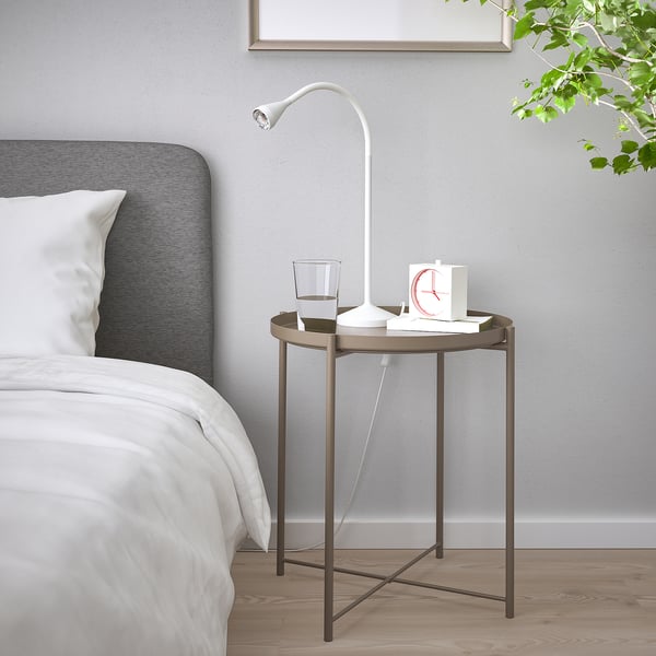 GLADOM black side table with white lamp, glass, clock, beside neatly made bed with white sheets, grey headboard, and wooden floor.