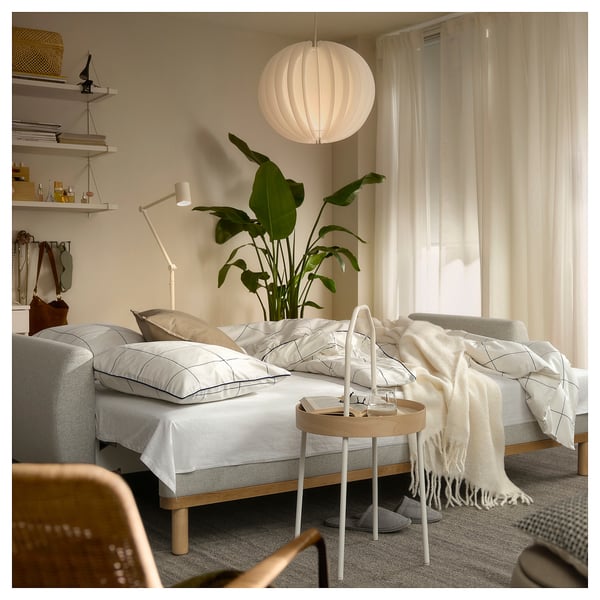 Well-lit room with white GRUNNARP sofa-bed, beige cushions, wooden side table, large plant, and bookshelf with accessories.