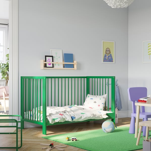 Kids room with green GULLIVER cot, wall shelves, toys, rug, and chair.
