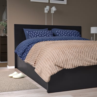 Neat bed with dark blue duvet, beige fleece, black headboard, slippers, and lamp.