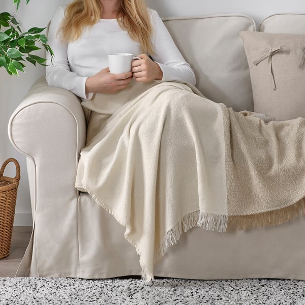 Person in white on couch, holding mug, under fringe throw, near plant.