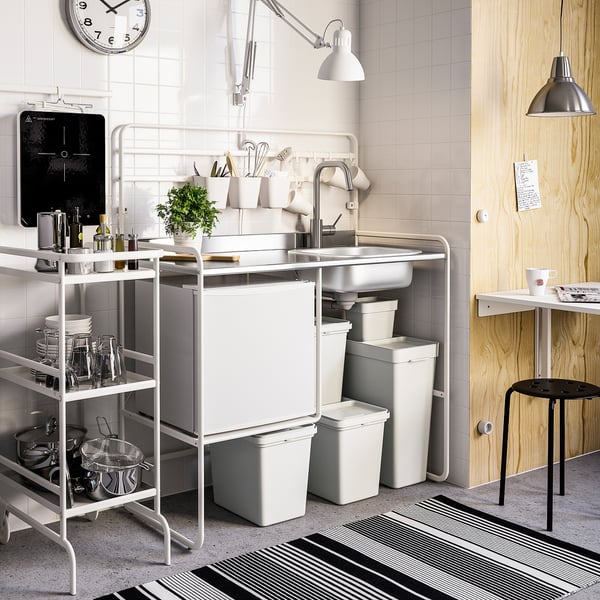 Tidy kitchen with white tiles, clock, shelves, and HÅLLBAR bins beside sink with dishes and plant.