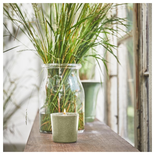 A lit green HEDERSAM scented candle sits beside a jar with grassy plants on a wooden shelf.
