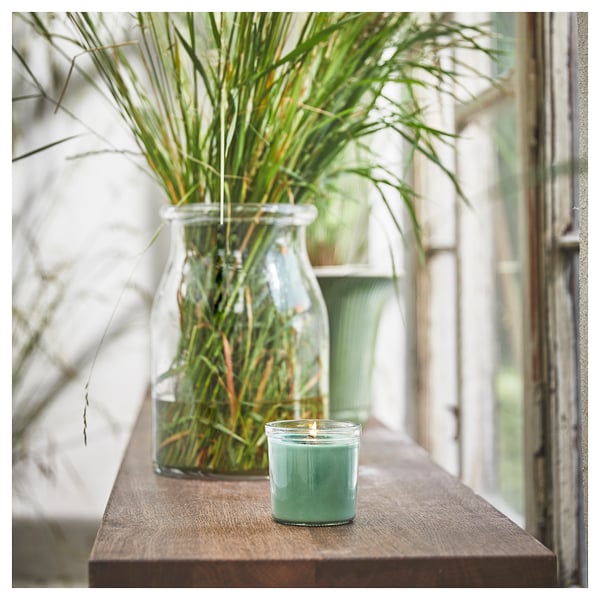 A lit teal green HEDERSAM scented candle sits beside a clear jar with greenery on a wooden shelf near a window.