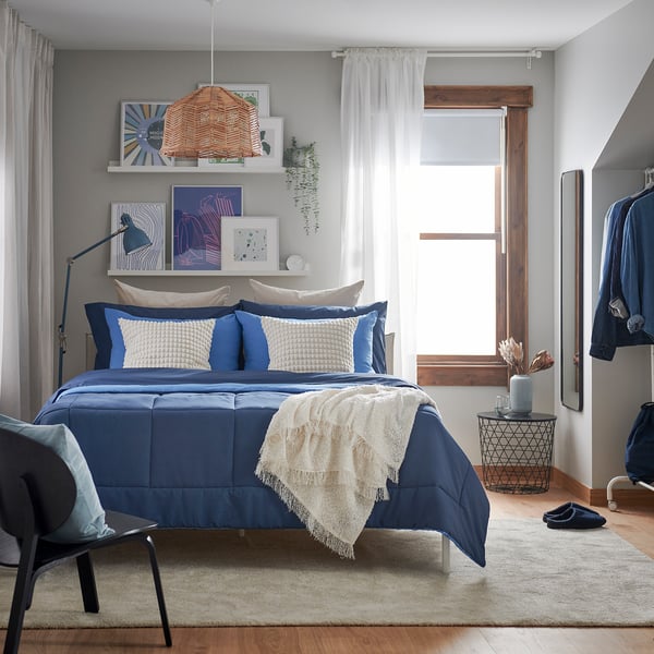 Bedroom with blue comforter set, white & blue pillows, art walls, large window, and spacious layout.