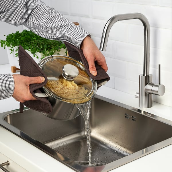 Person draining pasta in sink with gloves and apron, planter nearby.