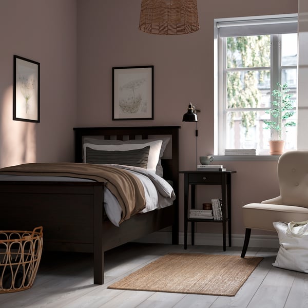 Cosy bedroom with HEMNES bed, rattan chair, & side table. Light purple walls, dark brown bed, beige chair, jute rug, & wall art.