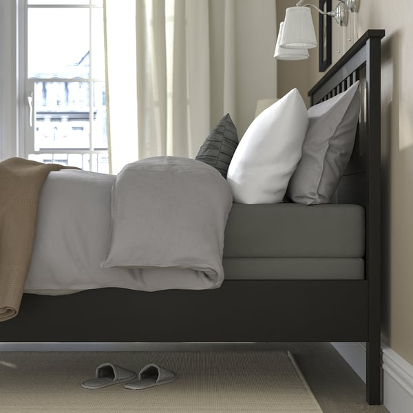 Bedroom scene: grey bedding on black wooden bed, against window with view to building. Two grey slippers below bed.