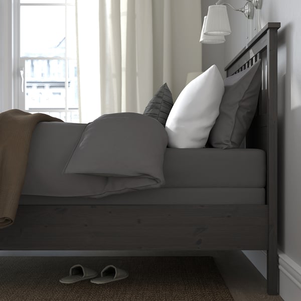 Bedroom with dark grey HEMNES bed and white pillows. Slippers under the bed; wall lamp nearby. Large window with sheer curtains.