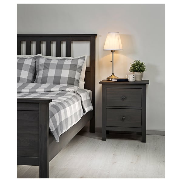 Bedroom scene: gray-checked bedding, wooden bed frame, bedside table with lamp, plant, and mug.
