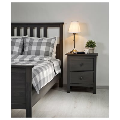 Bedroom scene: gray-checked bedding, wooden bed frame, bedside table with lamp, plant, and mug.