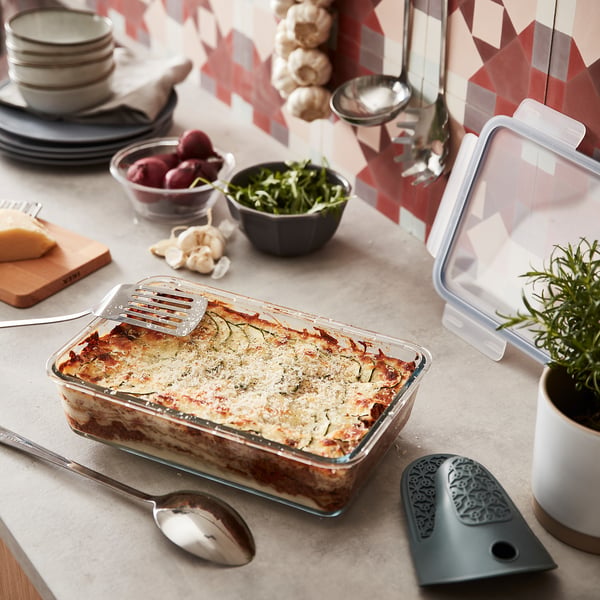 IKEA 365+ glass container holds a lasagna, larger than surrounding bowls, with utensils and plant nearby.
