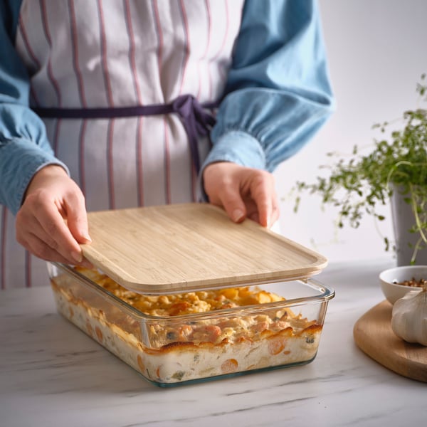 A person placing a bamboo lid on a glass baking dish filled with lasagna, demonstrating its use as a food cover.