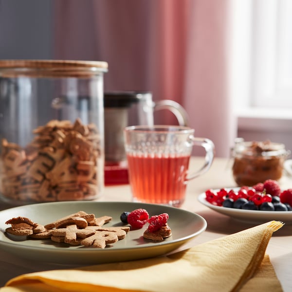 A breakfast table with KAFFEREP letter biscuits on a green plate, fresh berries, and a glass mug of red tea, set on a yellow cloth.