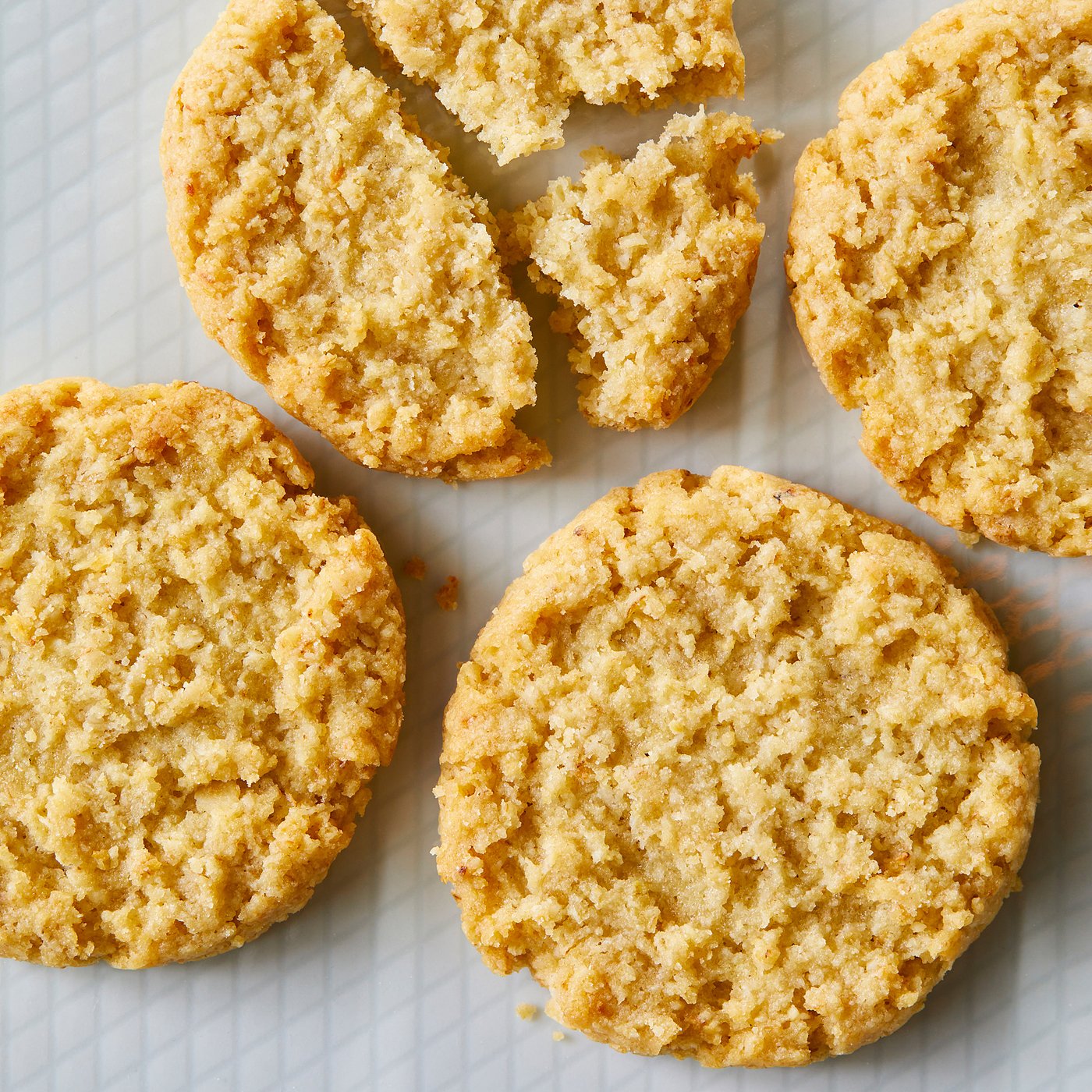 Six round, golden-brown oat biscuits on a white surface. One is broken, showing crumbly texture.